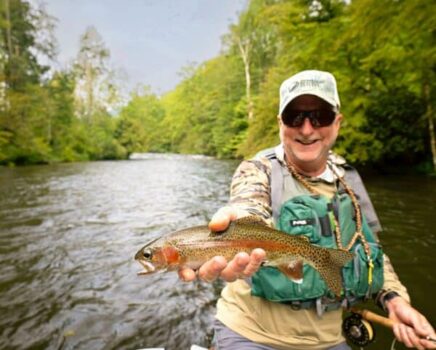 A man in fly fishing gear holds a trout with a river flowing in the background