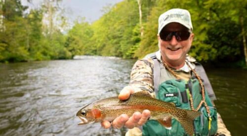 A man in fly fishing gear holds a trout with a river flowing in the background
