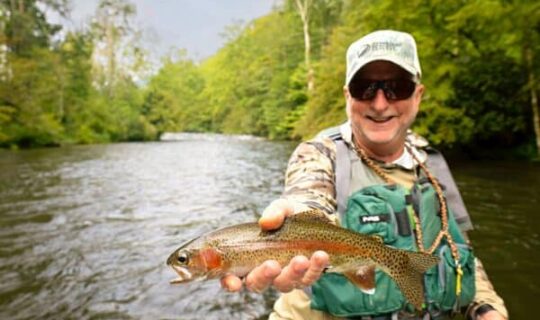 A man in fly fishing gear holds a trout with a river flowing in the background