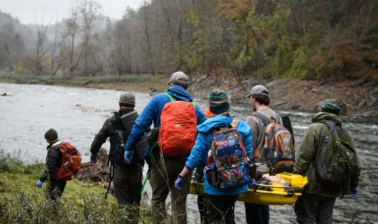 Group of people carrying a person in a stretcher by a river on the AWFA-WFR Bridge Course