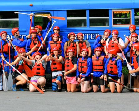 Guests in life jackets by a blue bus with paddles and they are all wearing a pfd. They all look happy and excited to be white water rafting.