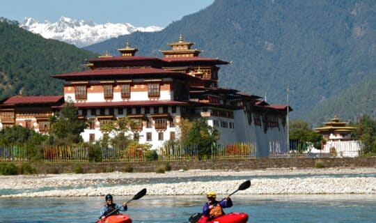 kayakers paddling in a river just below a colorful brown and white temple.