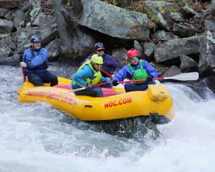 four people in a yellow rafting in rapids with rocks in the background