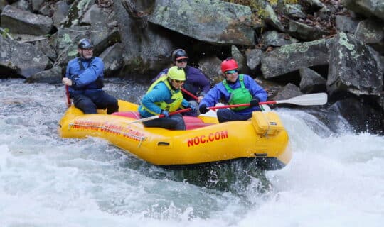 four people in a yellow rafting in rapids with rocks in the background