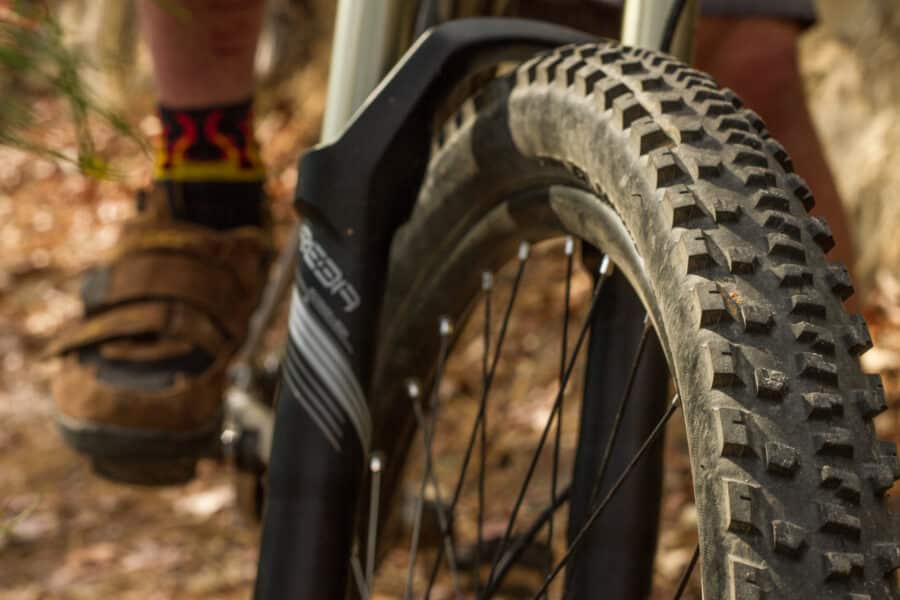A photo showing an up close image of a mountain bike tire, with a riders foot in the distance.