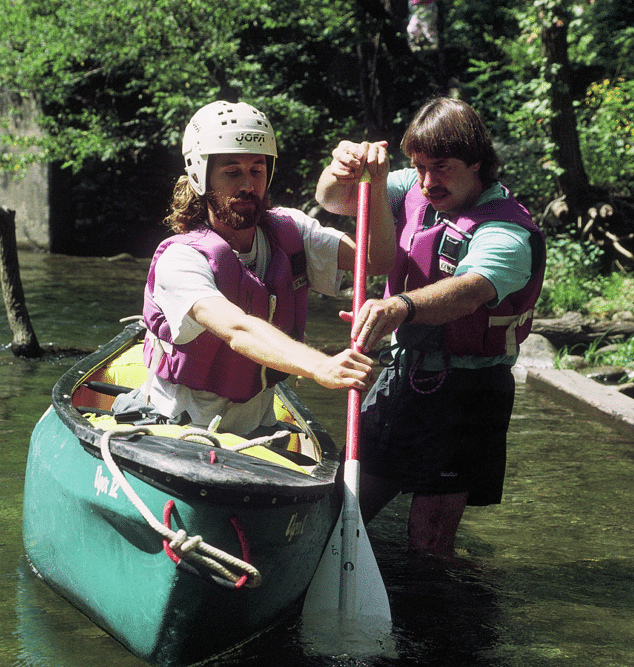 An instructor standing waist deep in water holds a canoe paddle with a man sitting in a green canoe.