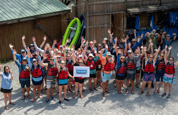 Large group of rafters in red life jackets raising their hands and smiling before a whitewater trip at NOC.