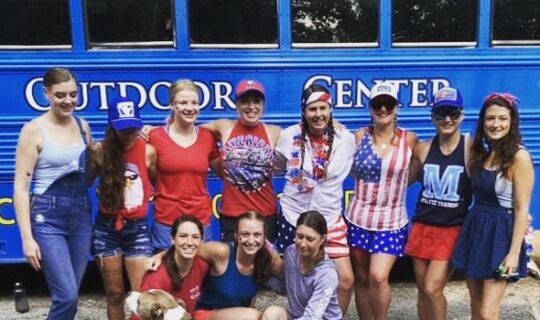A group of guests in America-themed outfits at the Nantahala Outdoor Center pose in front of a rafting bus with a dog.