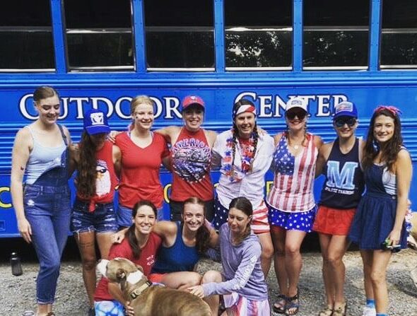 A group of guests in America-themed outfits at the Nantahala Outdoor Center pose in front of a rafting bus with a dog.