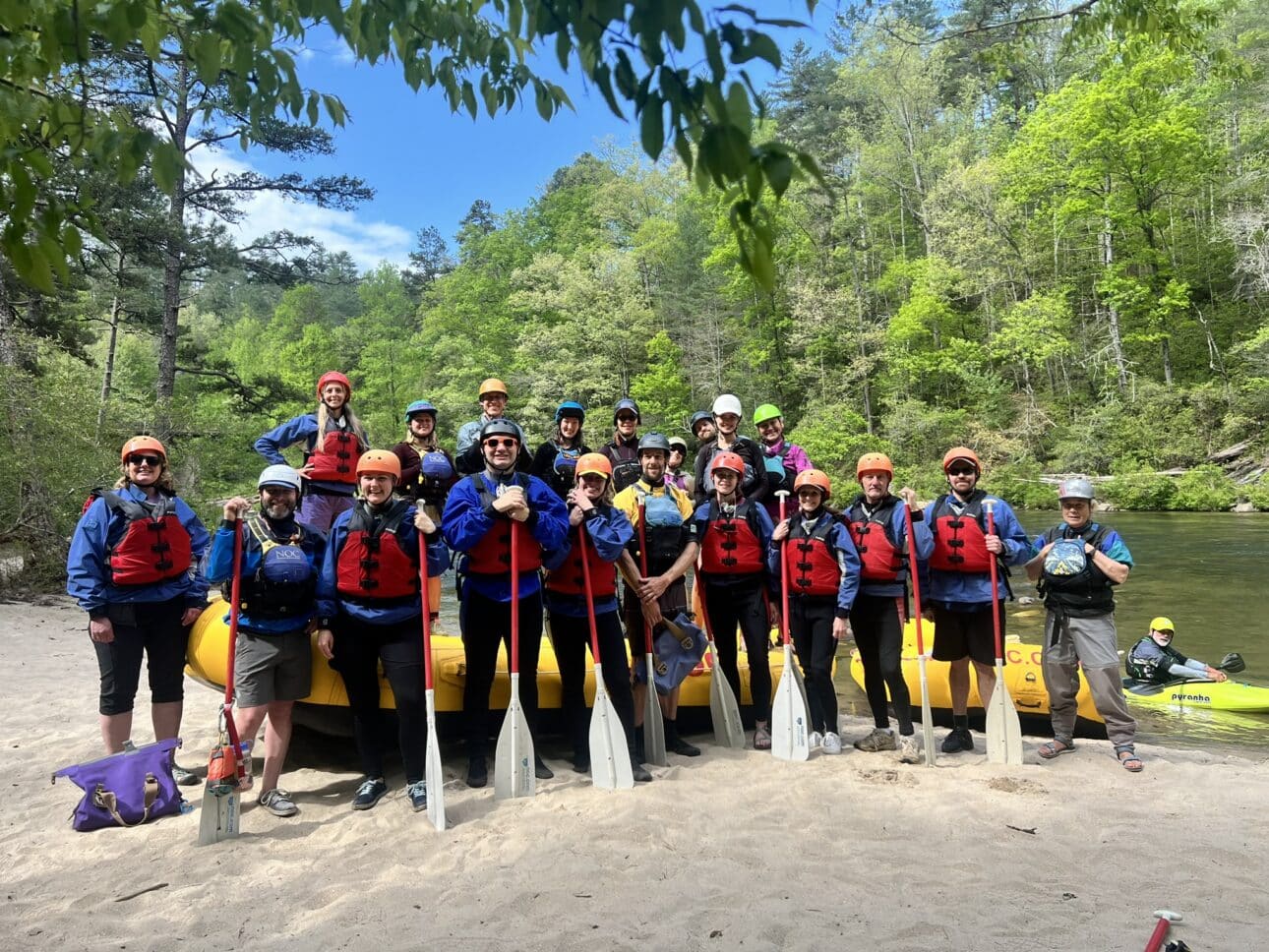 A group of rafting stand on a beach short with red pfds on, and paddles in their hands