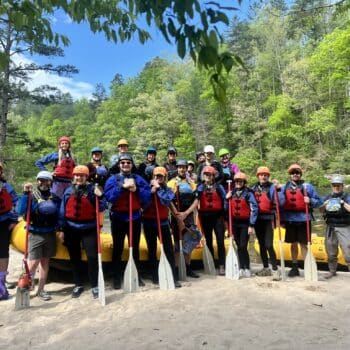 A group of rafting stand on a beach short with red pfds on, and paddles in their hands